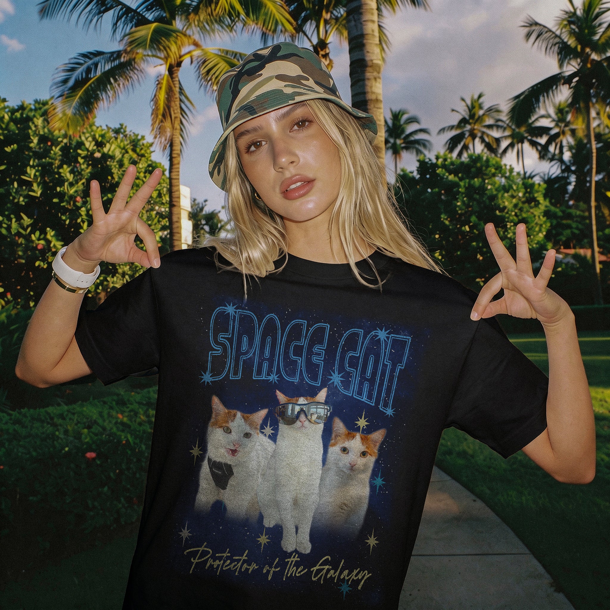 Image of A woman posing outdoors in front of palm trees, wearing a black graphic t-shirt with a 'SPACE CAT' design featuring three cats in space-themed artwork, and a camouflage bucket hat.